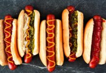 Hot Dog: Sokak Lezzetlerinin Vazgeçilmezi Dünya mutfağında geniş bir yer edinen hot dog, özellikle Amerika Birleşik Devletleri’nde adeta bir sembol haline gelmiştir. Kökeni 19. yüzyılın sonlarına kadar uzanan bu yiyecek, Almanya’dan Amerika’ya göç edenlerin beraberinde getirdiği sosis kültürü ile ortaya çıkmıştır. Uzun ekmek arasına yerleştirilen sosisin seneler içinde farklı soslarla buluşması, hot dog’u bugünkü popüler formuna kavuşturmuştur. Konulu bir haber görseli.
