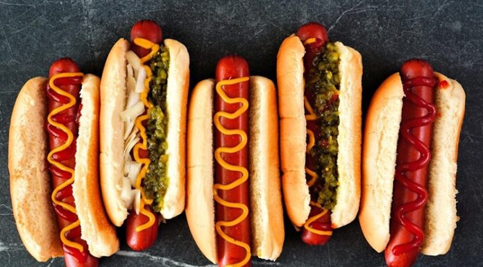 Hot Dog: Sokak Lezzetlerinin Vazgeçilmezi Dünya mutfağında geniş bir yer edinen hot dog, özellikle Amerika Birleşik Devletleri’nde adeta bir sembol haline gelmiştir. Kökeni 19. yüzyılın sonlarına kadar uzanan bu yiyecek, Almanya’dan Amerika’ya göç edenlerin beraberinde getirdiği sosis kültürü ile ortaya çıkmıştır. Uzun ekmek arasına yerleştirilen sosisin seneler içinde farklı soslarla buluşması, hot dog’u bugünkü popüler formuna kavuşturmuştur. Konulu bir haber görseli.