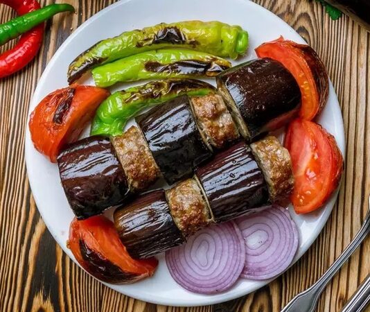 Patlıcan Kebabı: Sofraların Lezzetli ve Geleneksel Yemeği Türk mutfağının vazgeçilmez lezzetlerinden biri olan patlıcan kebabı, özellikle Gaziantep, Şanlıurfa ve Adana gibi şehirlerde sıkça hazırlanır. Patlıcanın közlenip etle buluşmasıyla ortaya çıkan bu yemek, hem hafif hem de doyurucu bir öğün sunar. Osmanlı mutfağından günümüze uzanan tarif, yöresel farklılıklarla her sofrada kendine özgü bir tat bırakır. Konulu bir haber görseli.